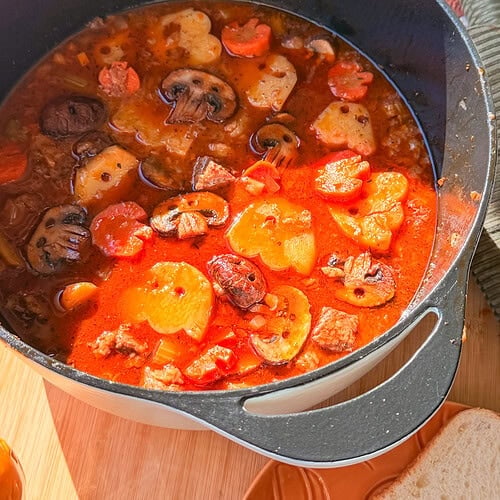 A black Dutch oven filled with a hearty stew containing mushrooms, carrots, potatoes, and chunks of meat in a tomato-based broth.