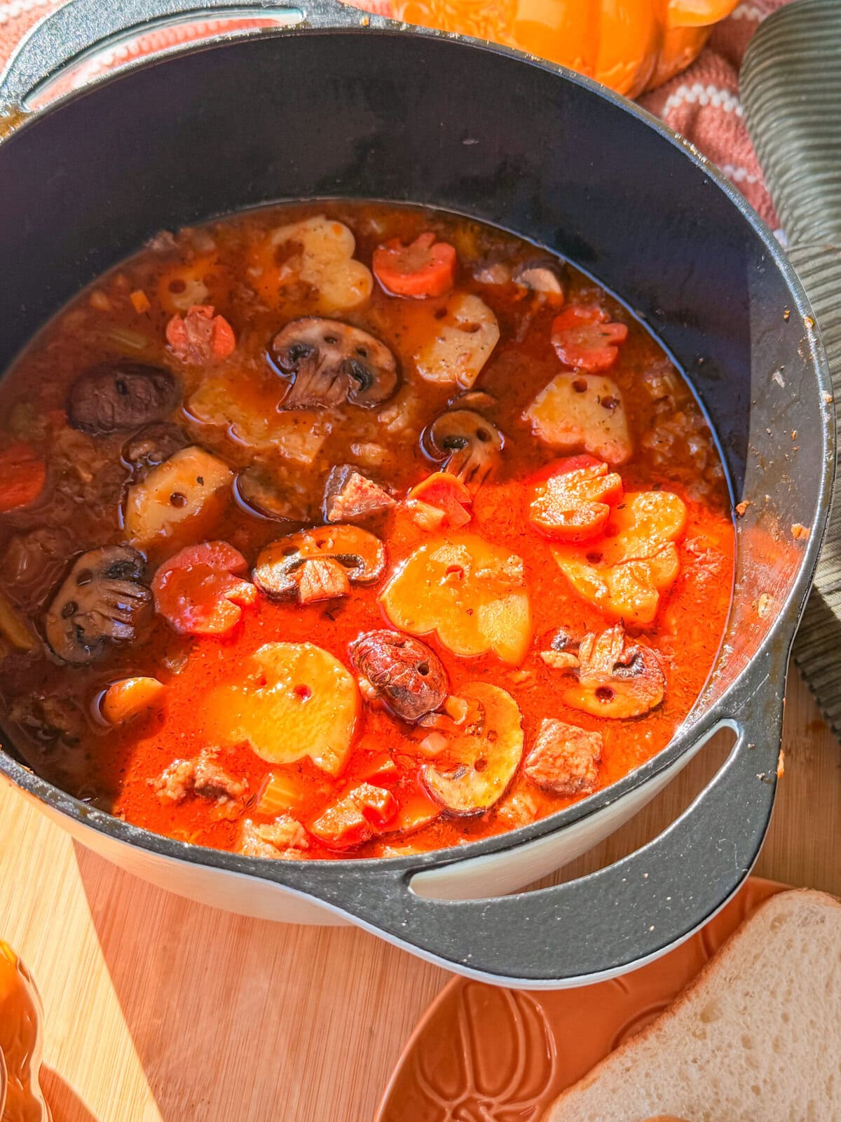 A black Dutch oven filled with a hearty stew containing mushrooms, carrots, potatoes, and chunks of meat in a tomato-based broth.