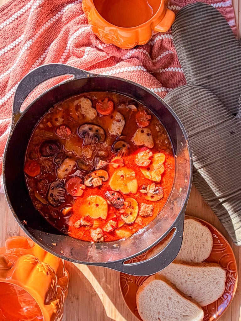 A cast iron pot filled with a hearty stew containing mushrooms and vegetables shaped like ghosts and pumpkins, served with slices of bread.