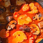 A hearty stew featuring chunks of potato, sliced mushrooms, and carrots in a vibrant red tomato-based broth.