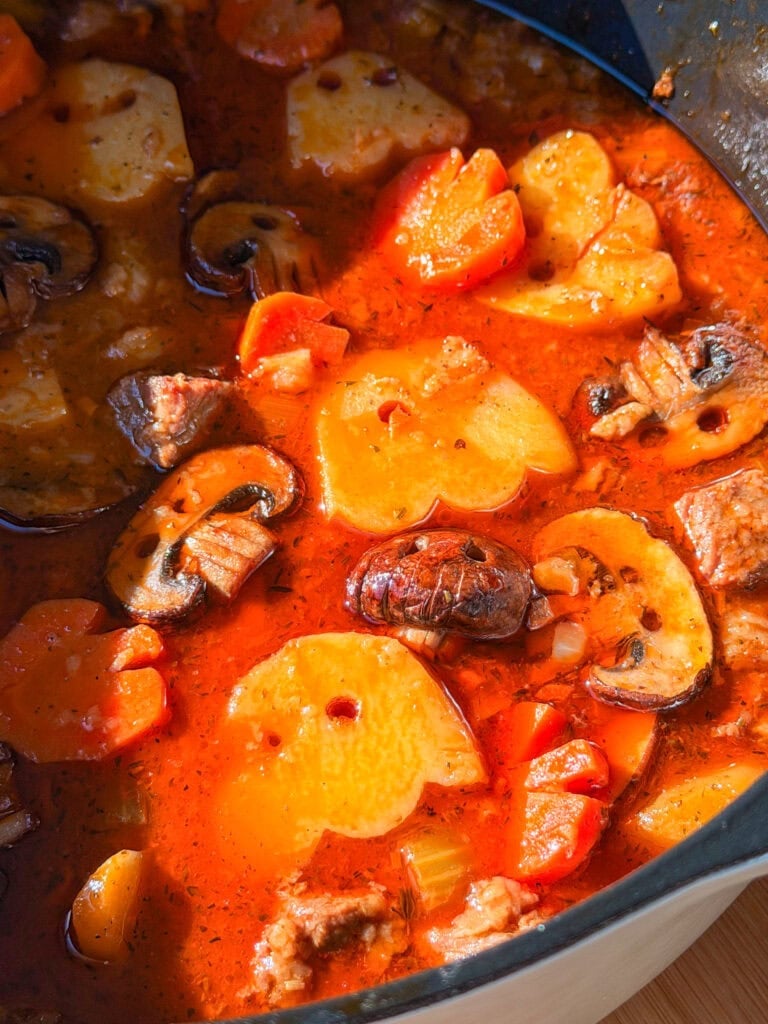 A hearty stew featuring chunks of potato, sliced mushrooms, and carrots in a vibrant red tomato-based broth.