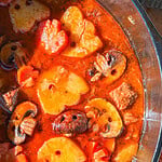 A pot of beef stew featuring chunks of meat, sliced mushrooms, and carrots in a rich red broth.