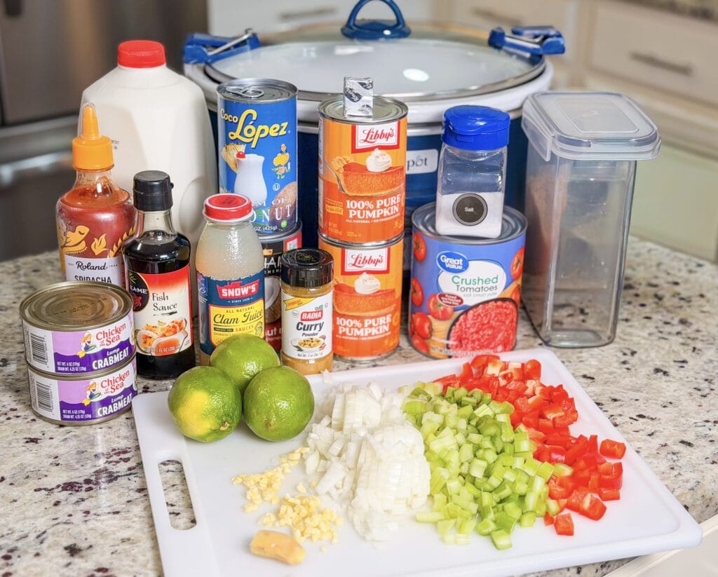 A collection of pantry staples including cans of crab meat, pumpkin puree, crushed tomatoes, coconut cream, clam juice, spices, sriracha, fish sauce, and a cutting board with chopped onion, celery, red bell pepper, and garlic.