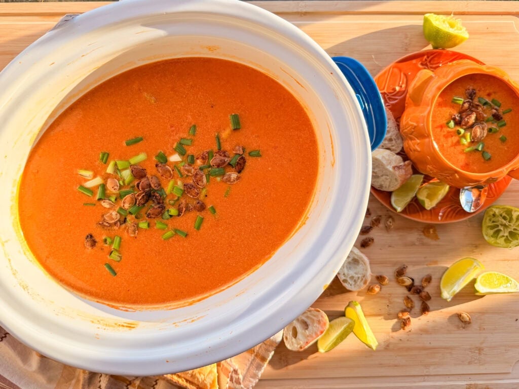 A large bowl and a smaller serving dish of creamy tomato soup topped with toasted seeds and chopped chives, accompanied by lime wedges and bread slices on a wooden surface.