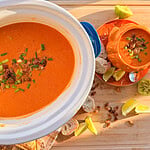 A large bowl and a smaller serving dish of creamy tomato soup topped with toasted seeds and chopped chives, accompanied by lime wedges and bread slices on a wooden surface.