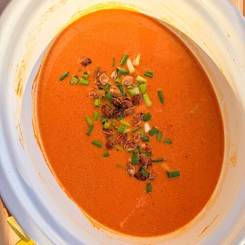 A slow cooker filled with creamy orange soup topped with toasted pumpkin seeds and chopped green onions.