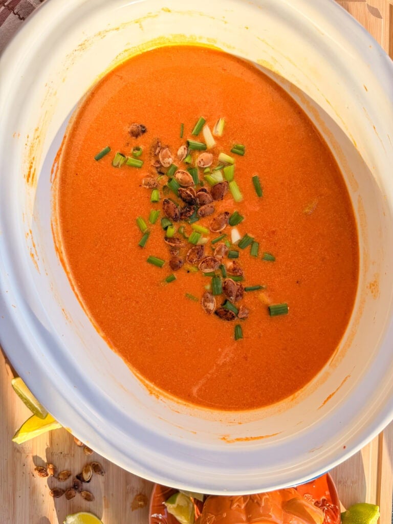 A slow cooker filled with creamy orange soup topped with toasted pumpkin seeds and chopped green onions.