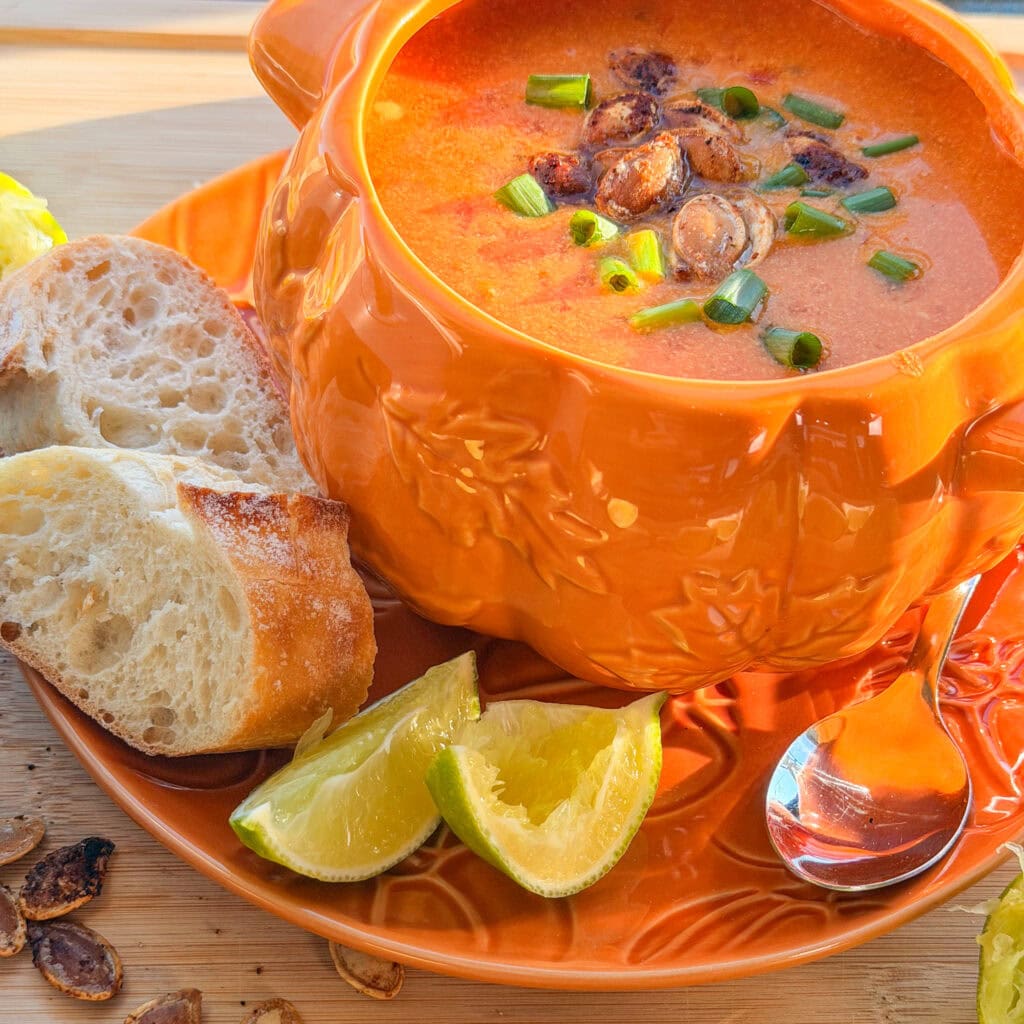 A pumpkin-shaped bowl filled with creamy tomato soup topped with roasted seeds and green onions, served with two slices of bread and lime wedges.