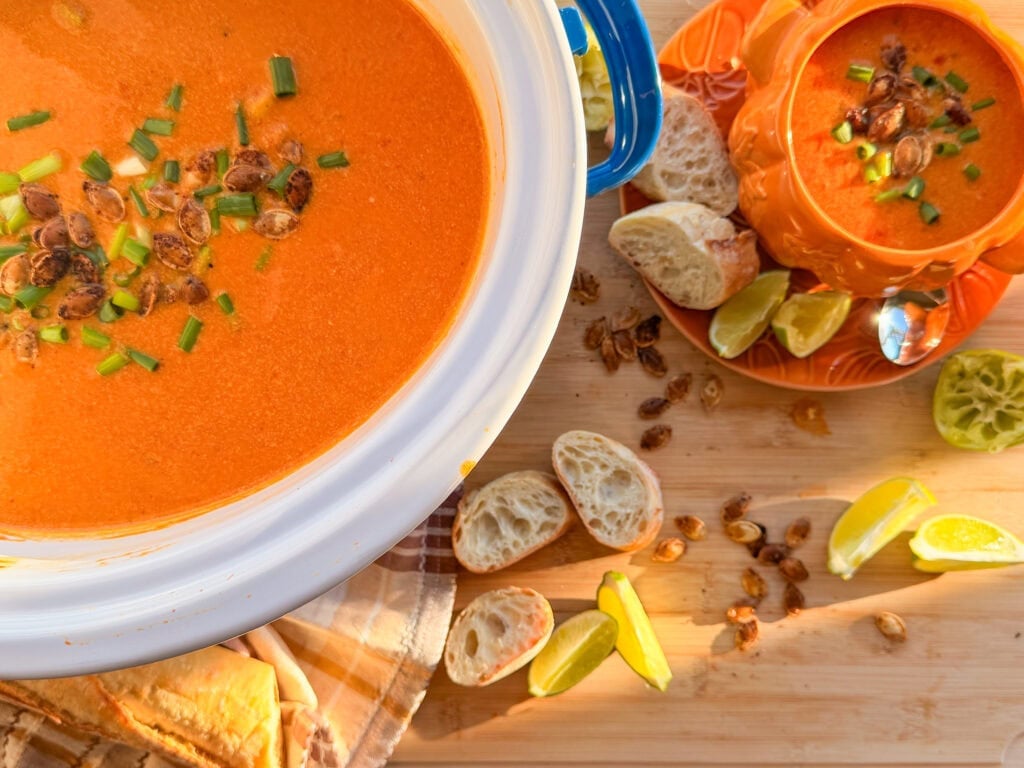 A large pot and a pumpkin-shaped bowl filled with tomato soup, topped with toasted pumpkin seeds and green onions, served with sliced baguette and lime wedges.