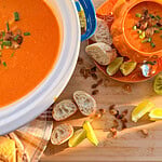 A large pot and a pumpkin-shaped bowl filled with tomato soup, topped with toasted pumpkin seeds and green onions, served with sliced baguette and lime wedges.