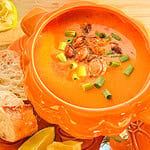 A bowl of creamy orange soup topped with roasted seeds and chopped green onions, served with lime wedges and slices of crusty bread.