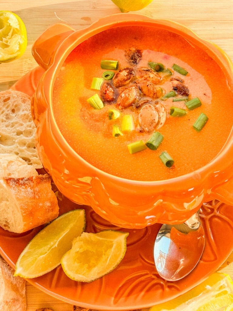 A bowl of creamy orange soup topped with roasted seeds and chopped green onions, served with lime wedges and slices of crusty bread.